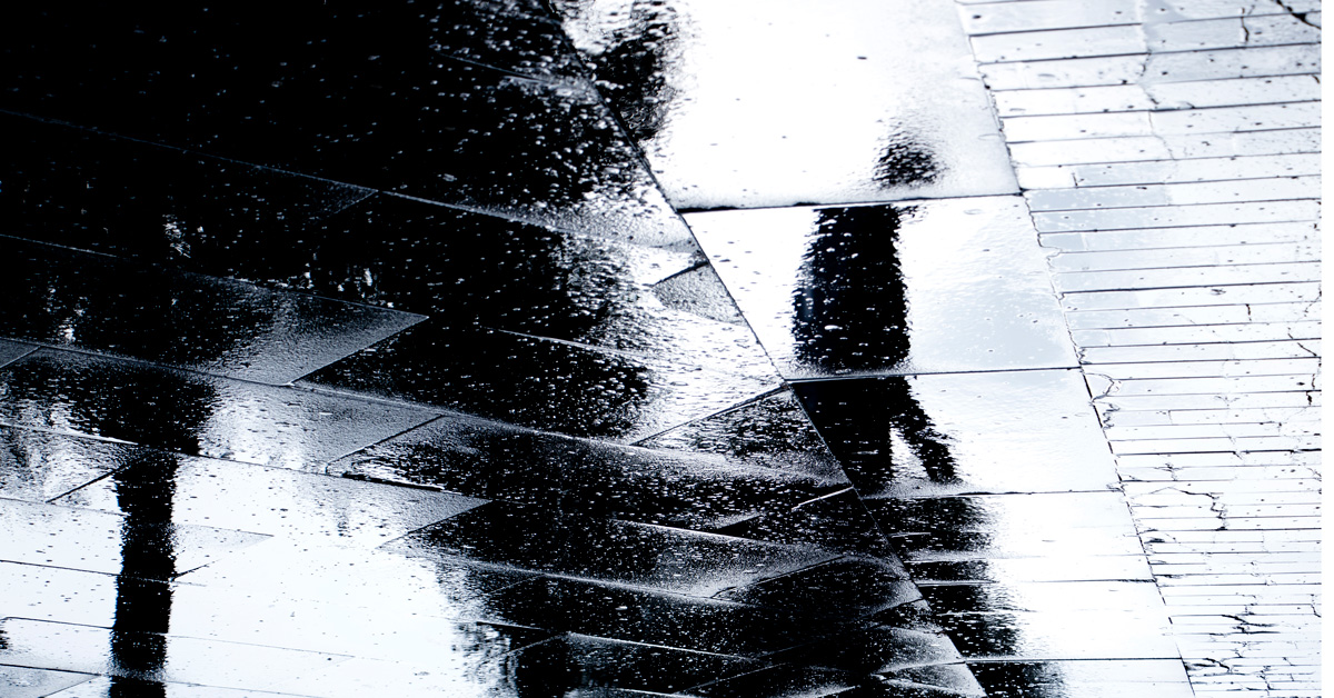 Asfaltista heijastuu yksinäisen kävelijän hahmo. Mustavalkoinen kuva.