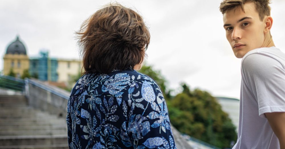 Poika istuu kaiteella ja katsoo kameraan vierellään nainen.